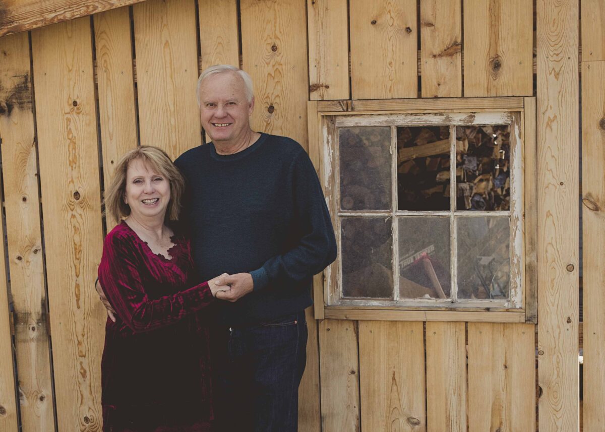 A man and a woman hold hands and smile in front of a cabin.