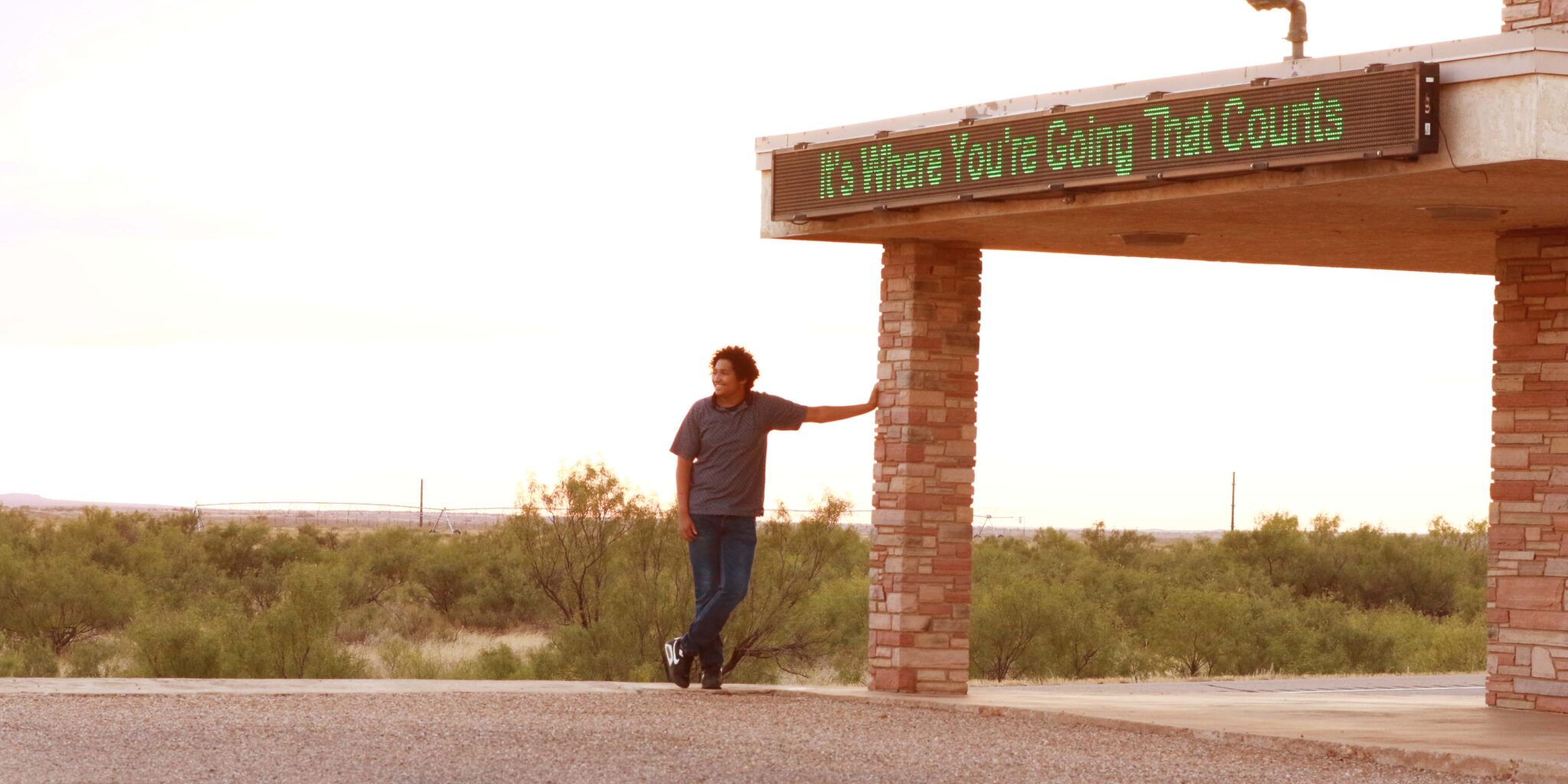 A boy leans on a post in front of the setting sun.