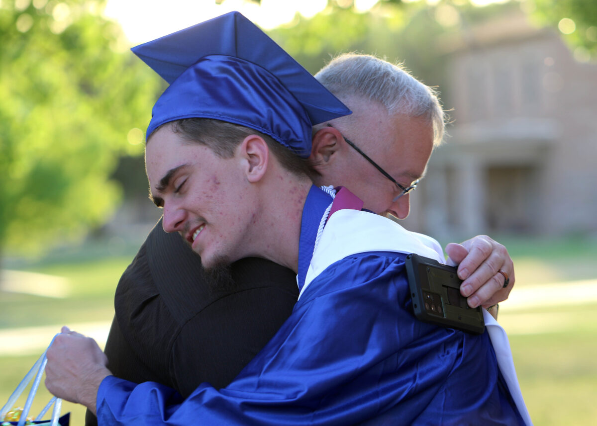 A teenager in a blue graduation cap and gown hugs an older man.
