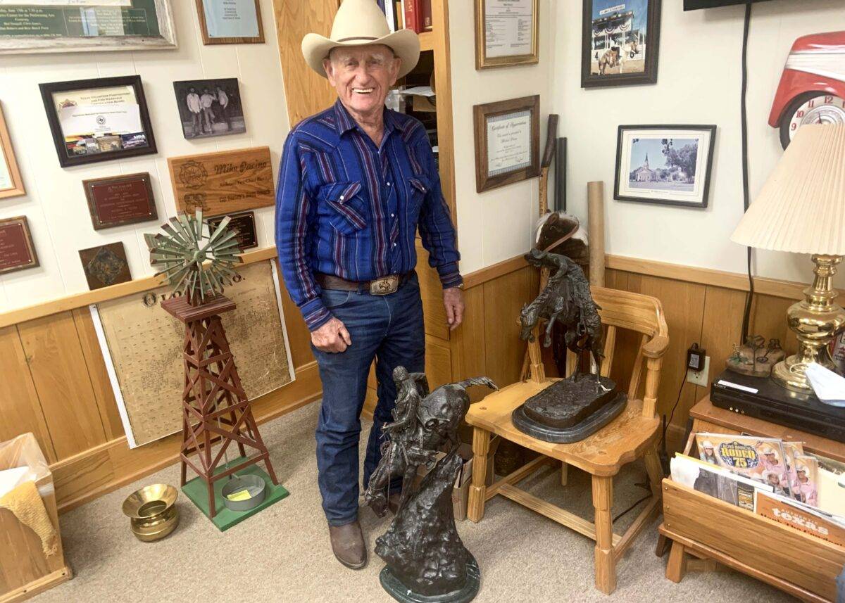 A man wearing a cowboy hat and a cowboy plaid shirt stands behind a western sculpture.