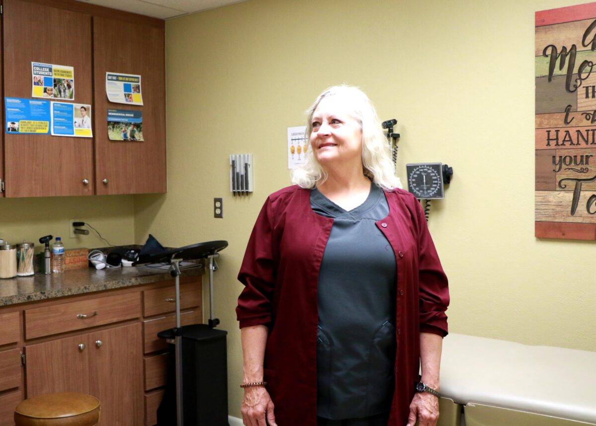 A silver-haired nurse wearing a maroon sweater stands in a doctor's office.